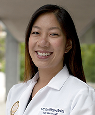 Smiling woman with long dark hair wearing a white lab coat standing outdoors.