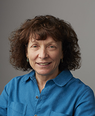 Portrait of a woman with curly brown hair wearing a blue shirt against a gray background.