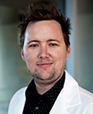 Portrait of a man with short spiked hair wearing a white coat over a black shirt, smiling slightly.