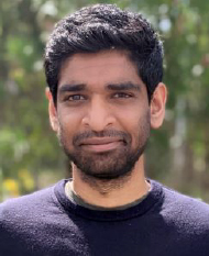 Man with short black hair and beard smiling outdoors with blurred green foliage background.