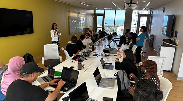 A diverse group of people in a conference room attending a presentation, with a woman standing and speaking at the front near a yellow wall and two large monitors.