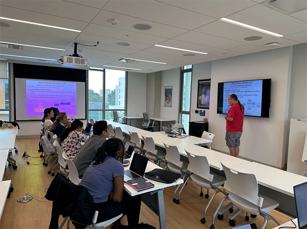 Classroom setting with a group of students seated and facing a presenter in a red shirt, who is standing near a wall-mounted screen displaying a presentation.