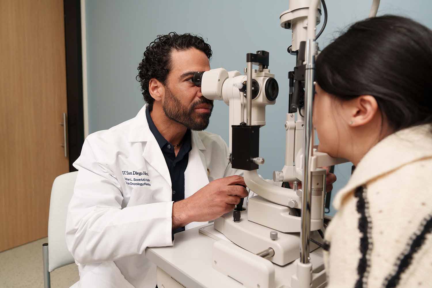 An eye doctor in a white coat conducts an eye exam using a slit lamp on a female patient seated opposite him.