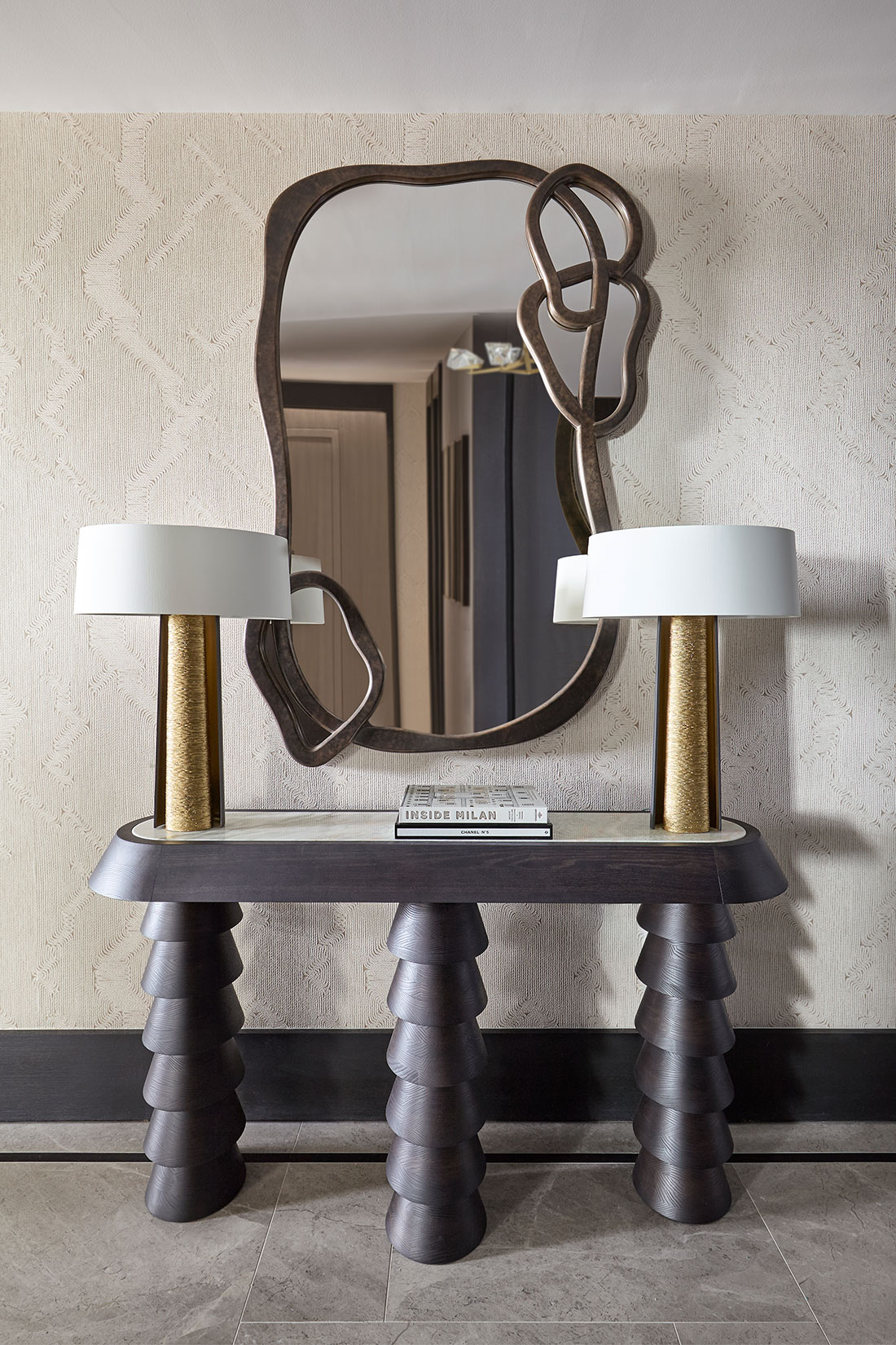 Textured wallpaper with contemporary timbre console table and pair of gold lamps. Above sits a abstract mirror in bronze metal finish.