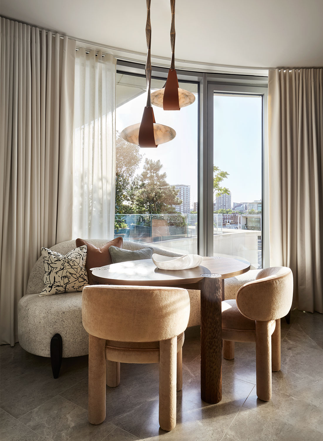 Curved kitchen sofa around wooden carved table. Above hangs alabaster lights in leather straps. In the backrgound the window provides view over London.