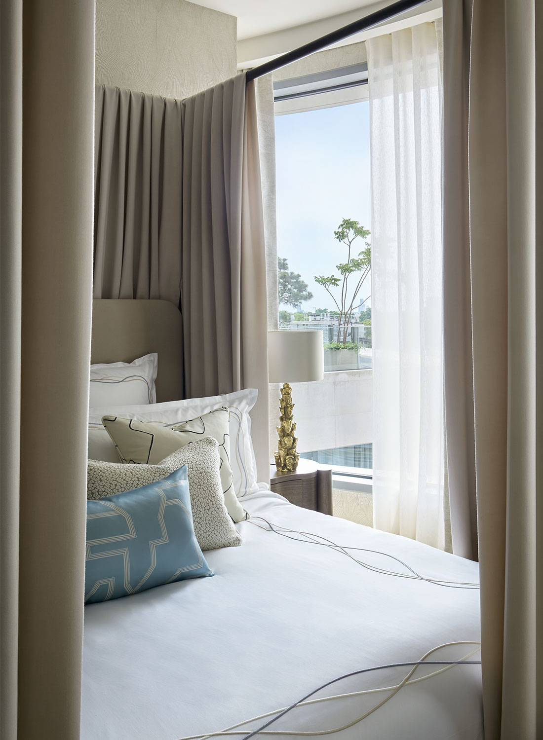 View through four poster bed silk curtain to reveal glimpse of luxury bed linen, bedside table and lam. Out of the window you can see view of London.