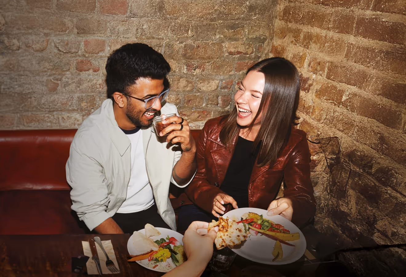 Two friends laughing and sharing food at a table with exposed brick walls in the background.