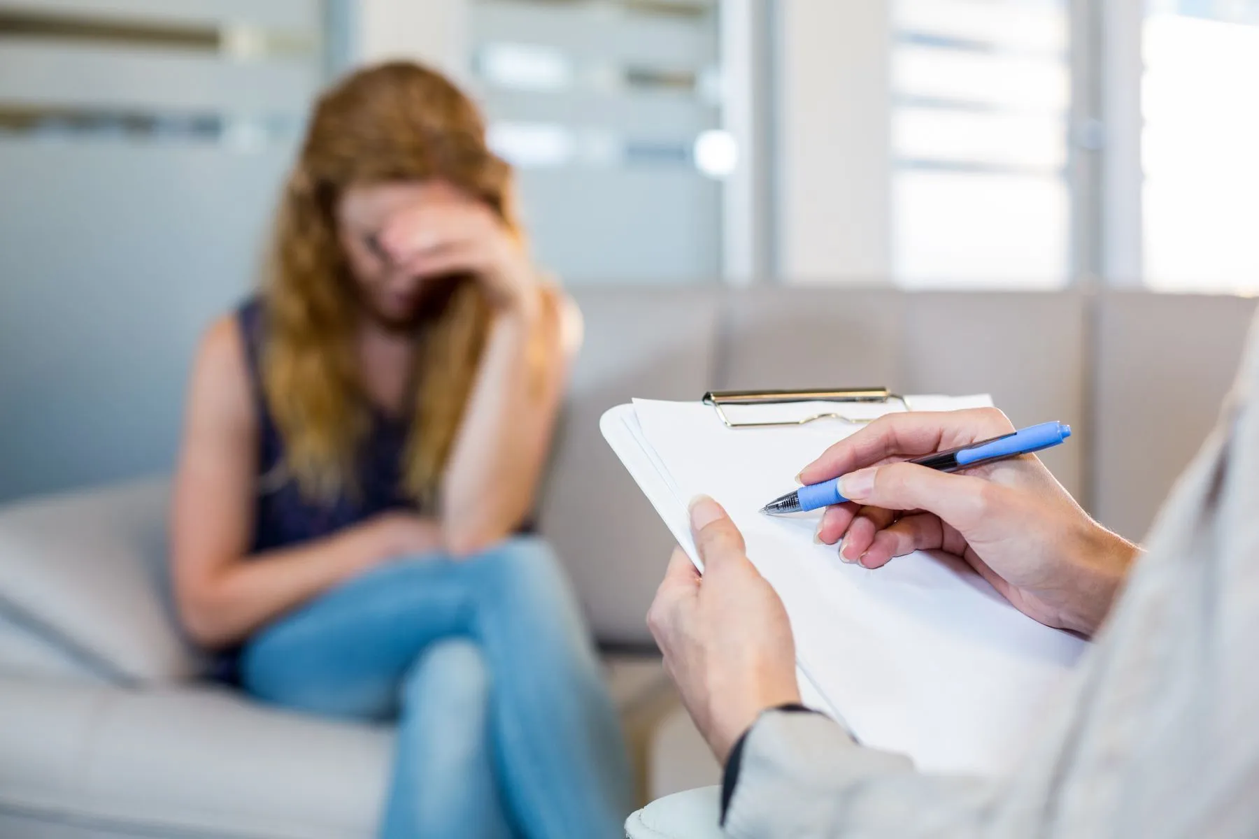 Young woman sitting on couch with hand across her face and therapist taking notes about depression.