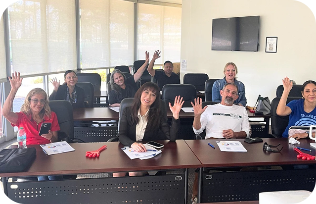 A group of seven people seated in a classroom raising one hand, with papers, phones, and water bottle on desks.