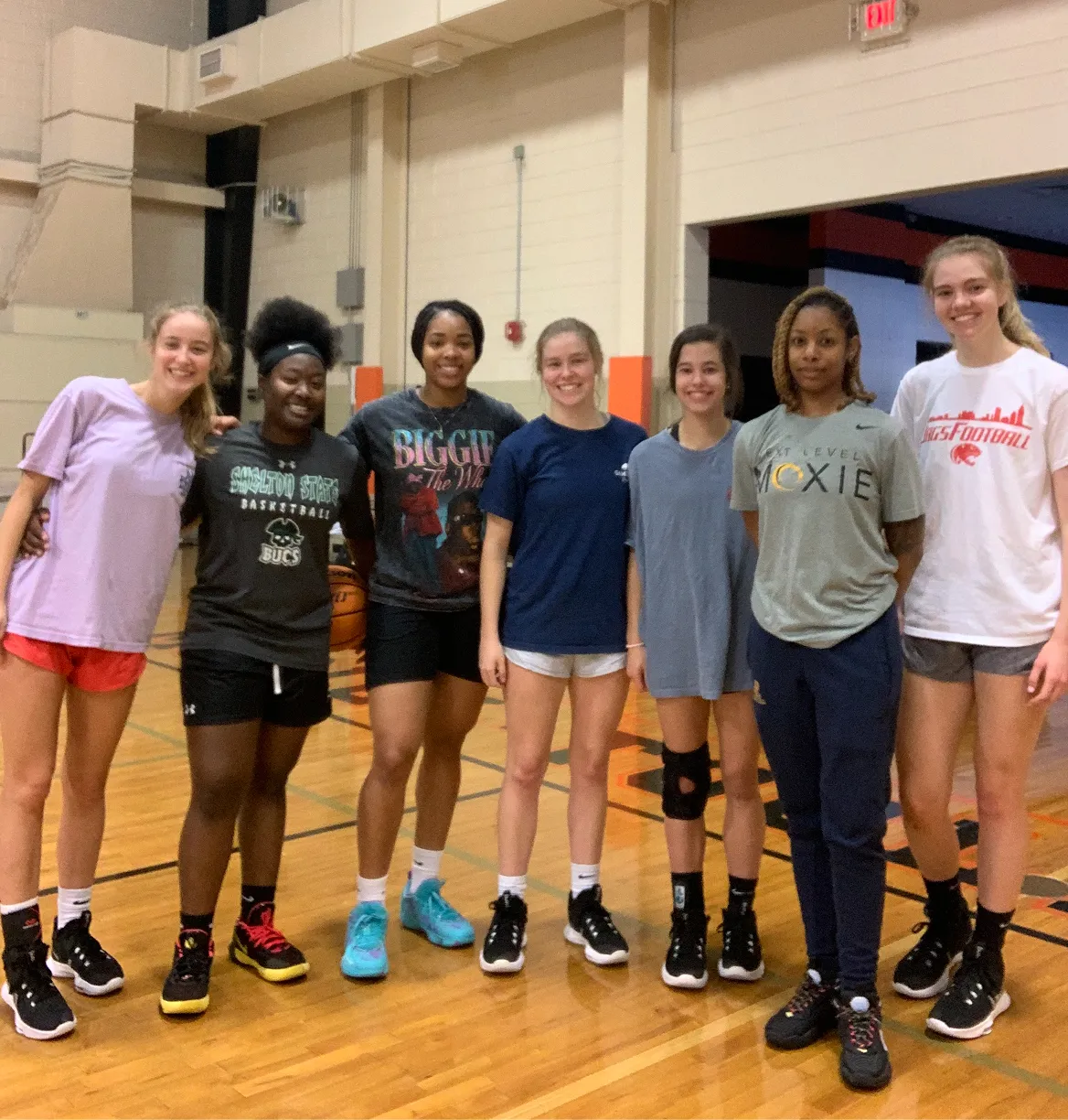 Seven young women standing in a gym basketball court wearing athletic clothing, smiling at the camera.