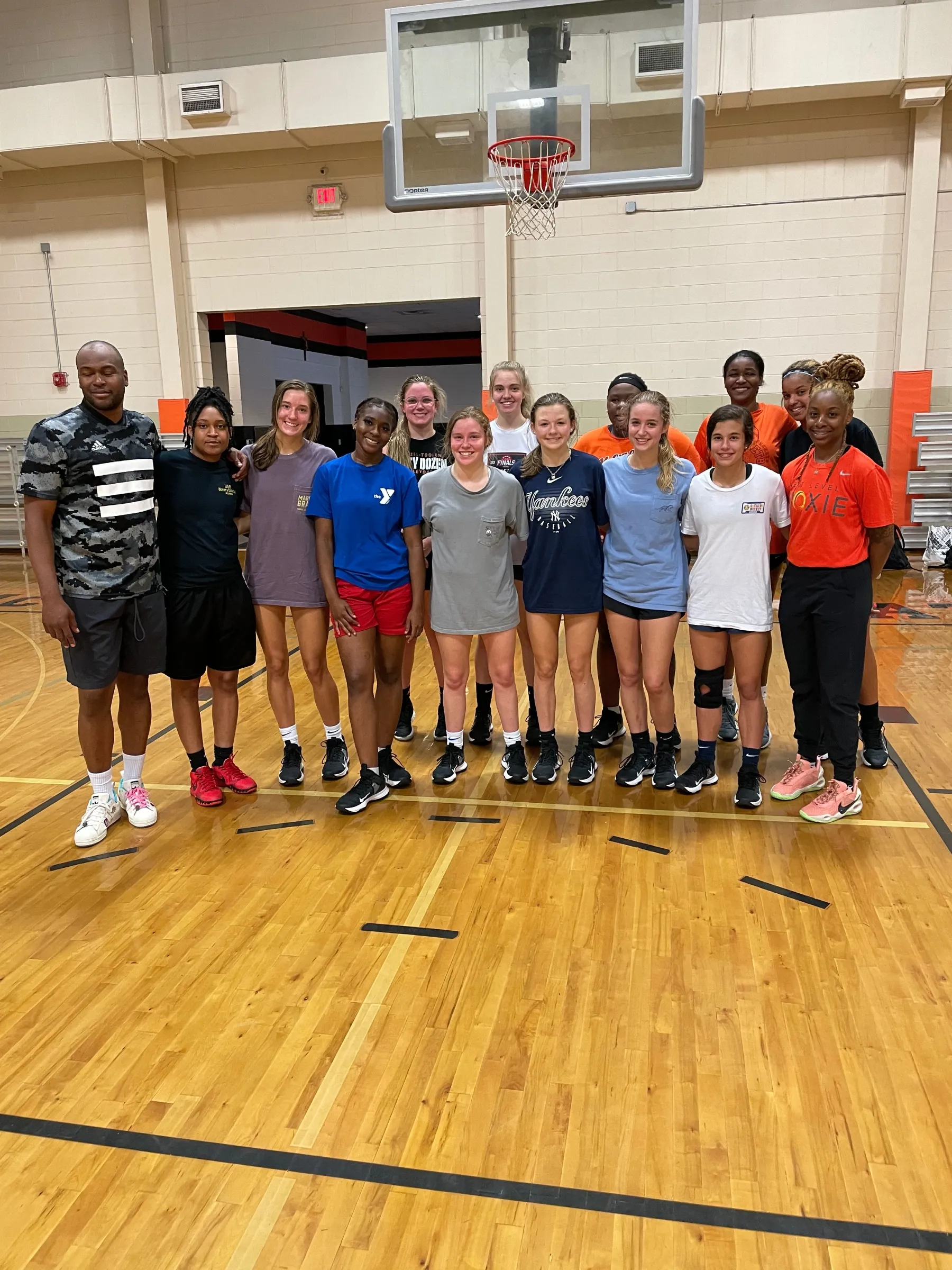 Seven young women standing in a gym basketball court wearing athletic clothing, smiling at the camera.