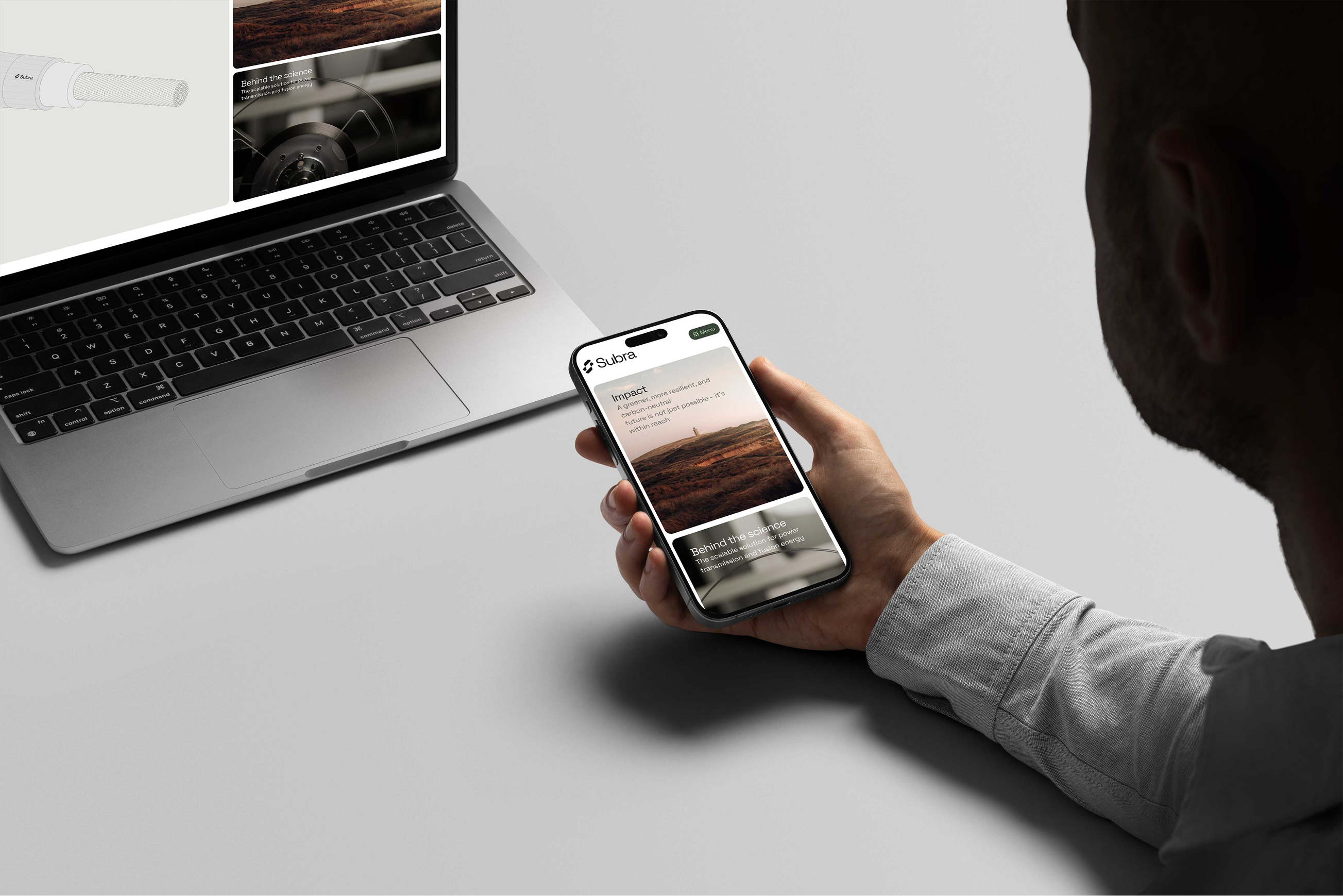Person holding smartphone displaying a website with text 'Impact' and 'Behind the science' next to a laptop on a gray desk.