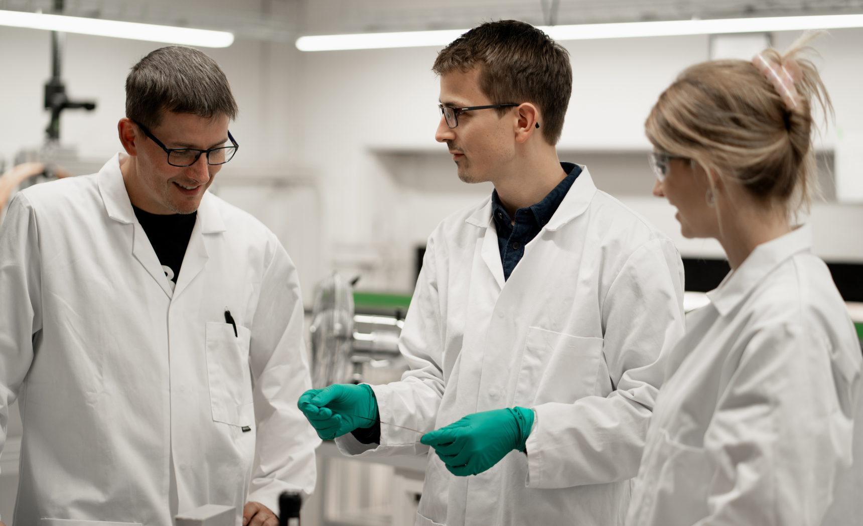 Three scientists in lab coats discussing and examining a thin object in a laboratory setting.