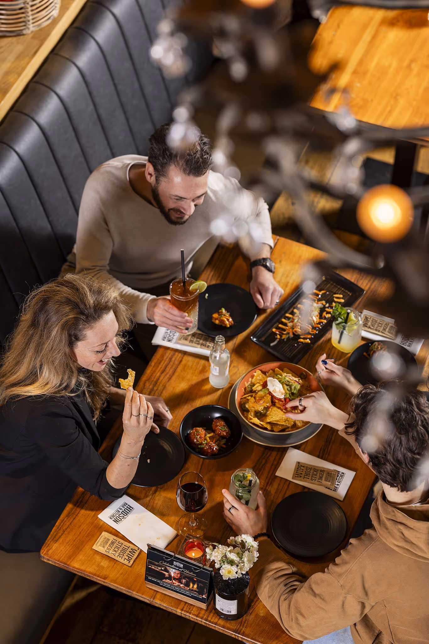 Drie gasten zitten en lachen aan een houten tafel gevuld met kleine gerechten.