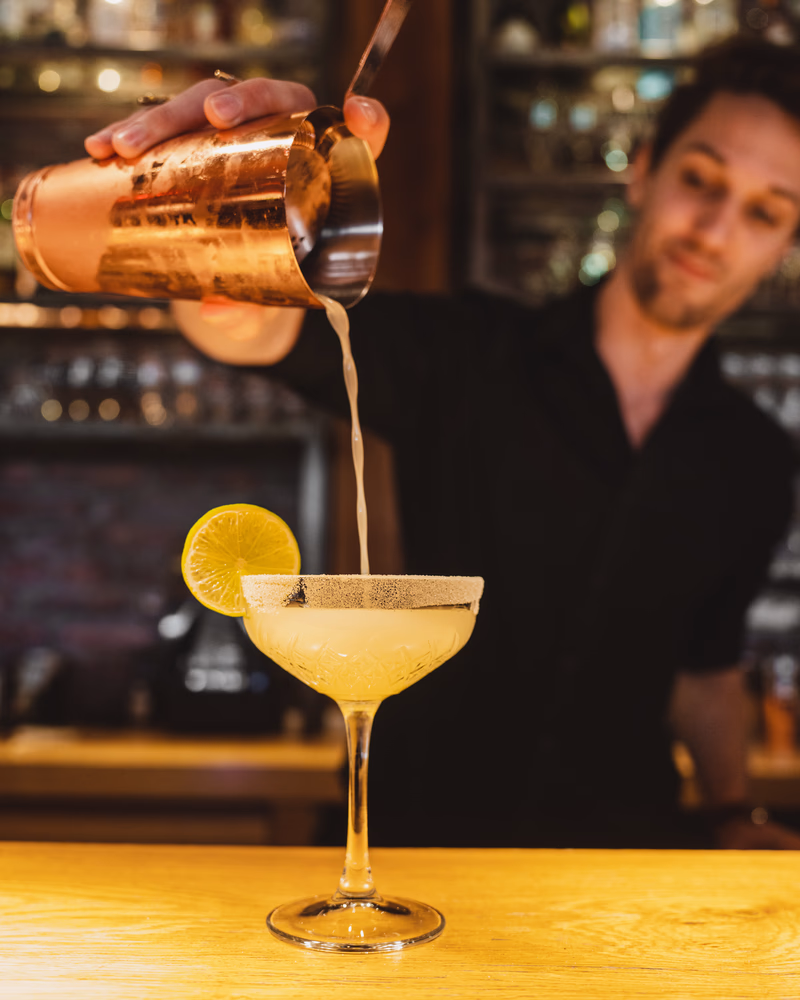 Barman schenkt een geelkleurige cocktail in een cocktailglas met een citroentje.