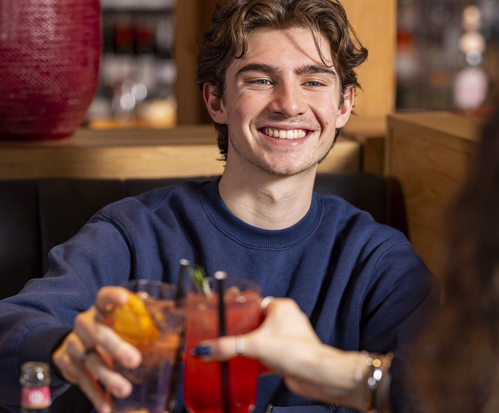 Lachende jonge man proost met een ander persoon aan een tafel met gerechten.