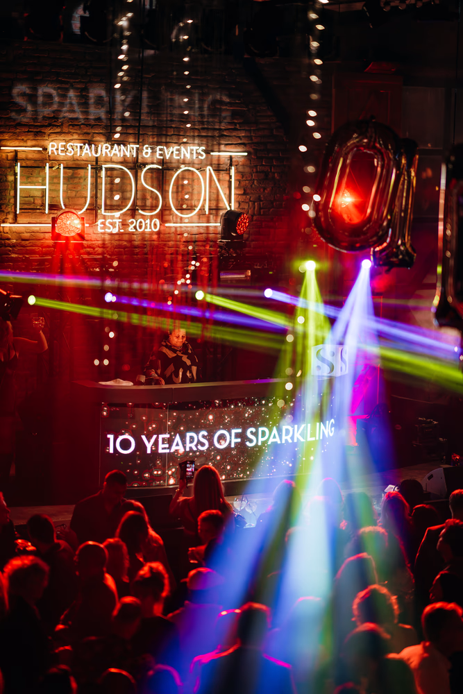 Drukke nachtclubsetting met een DJ achter de draaitafel en kleurrijke lichtstralen boven een feestend publiek.