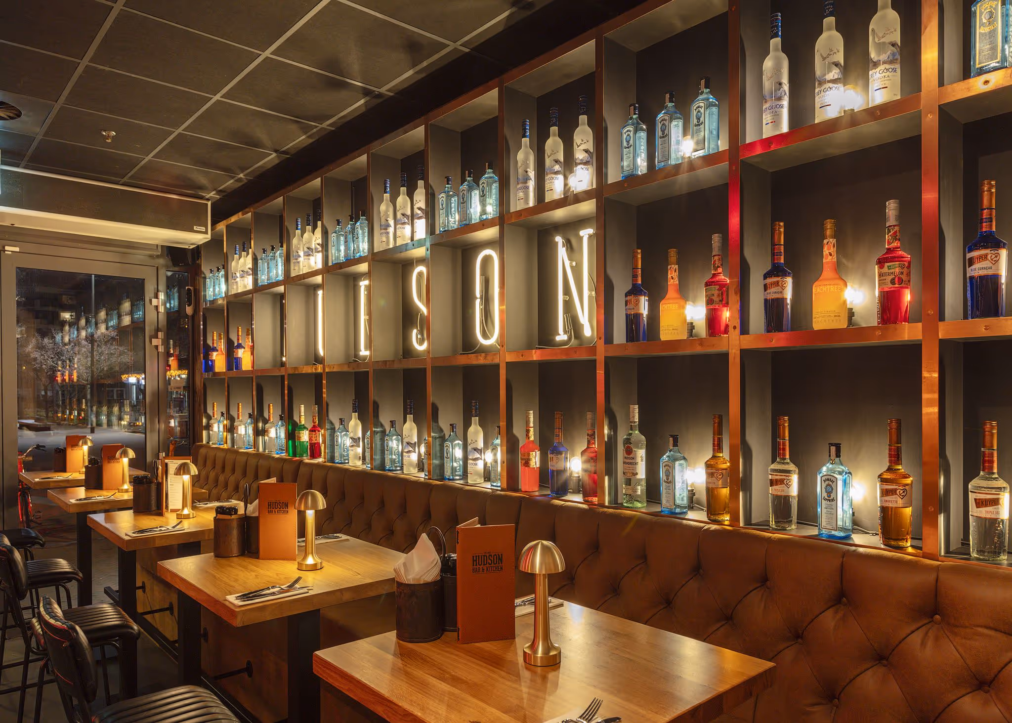 Gezellige bar en restaurant met houten tafels, wijnglazen en sfeerverlichting die de naam Hudson in rode letters toont.
