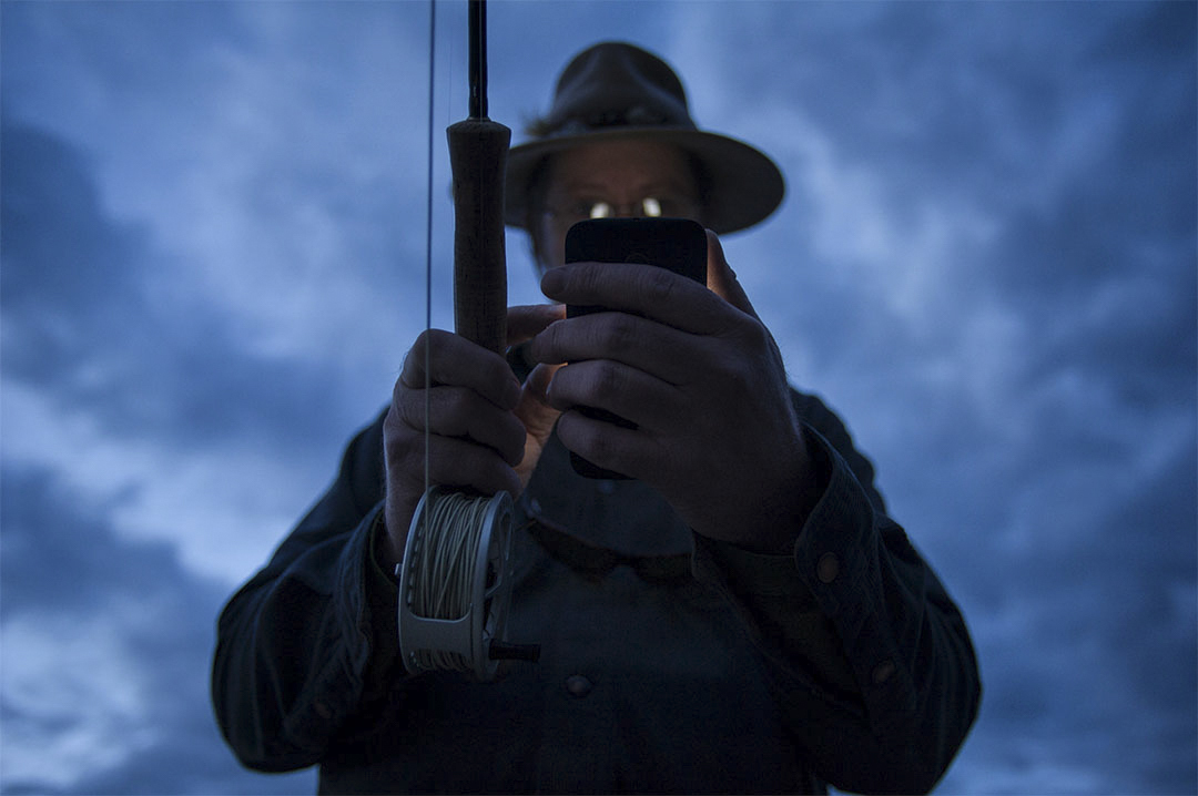 An old man with a fly rod looking at this cell phone.