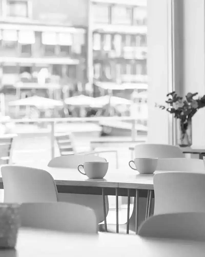 Black and white photo showing a café interior with empty chairs and tables, two cups on a table, and a window with a blurry outdoor view.