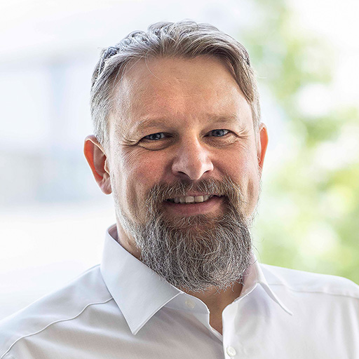 Smiling middle-aged man with grey hair and beard wearing a white shirt against a blurred outdoor background.