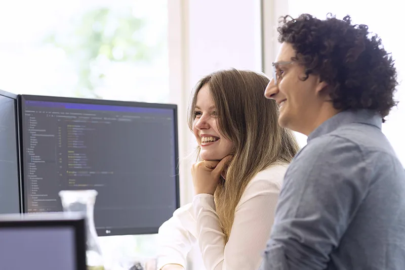 Two coworkers smiling and looking at a computer screen displaying code in a bright office.