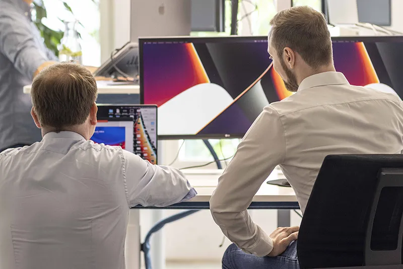 Two men in white shirts sitting at a desk looking at a laptop and a large monitor in a bright office.