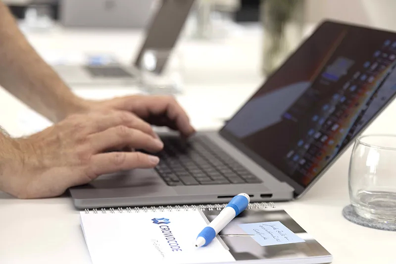 Person typing on a laptop keyboard with a clear glass, a blue and white pen, and a notebook labeled 'CrowdLogic' on a white desk.