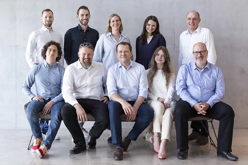 Group portrait of ten diverse professionals smiling and seated or standing against a plain gray wall.