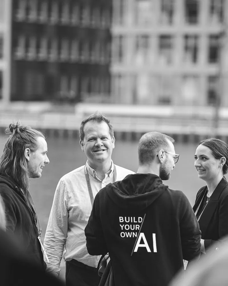 Four people engaged in conversation outdoors, with one person wearing a hoodie that says 'BUILD YOUR OWN AI'.