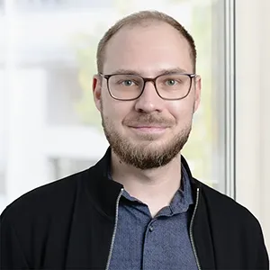 Smiling man with glasses, beard, and short hair wearing a dark jacket over a blue collared shirt, standing near a window.