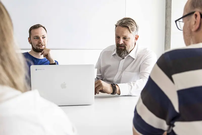 Four people in a meeting around a laptop, engaged in discussion in a bright room.