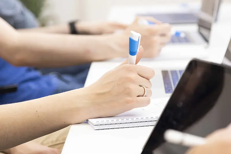 Hands holding pens, writing on notepads with laptops in the background, likely in a seminar or class.