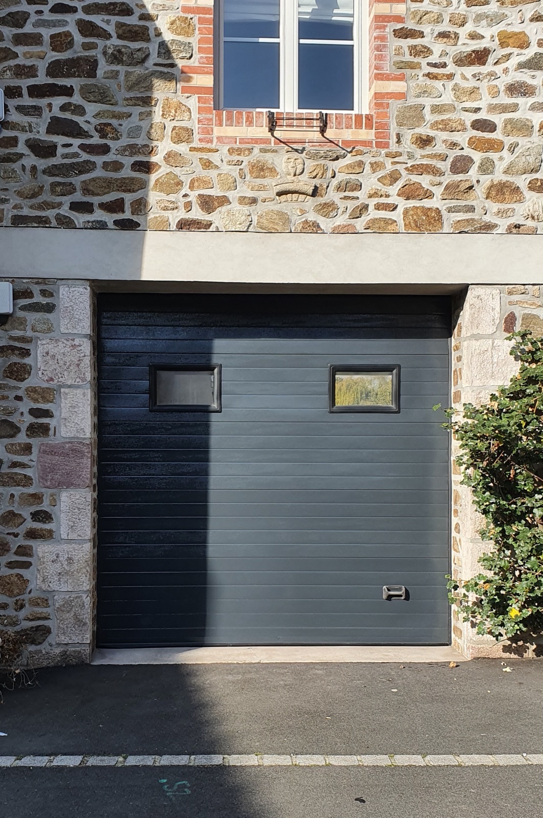 Porte de garage sectionnelle anthracite encadrée de pierre dans une façade en moellons