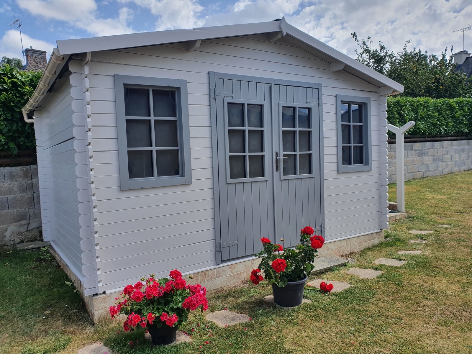 Abri de jardin en bois peint en blanc, porte et fenêtres gris clair, pots de fleurs