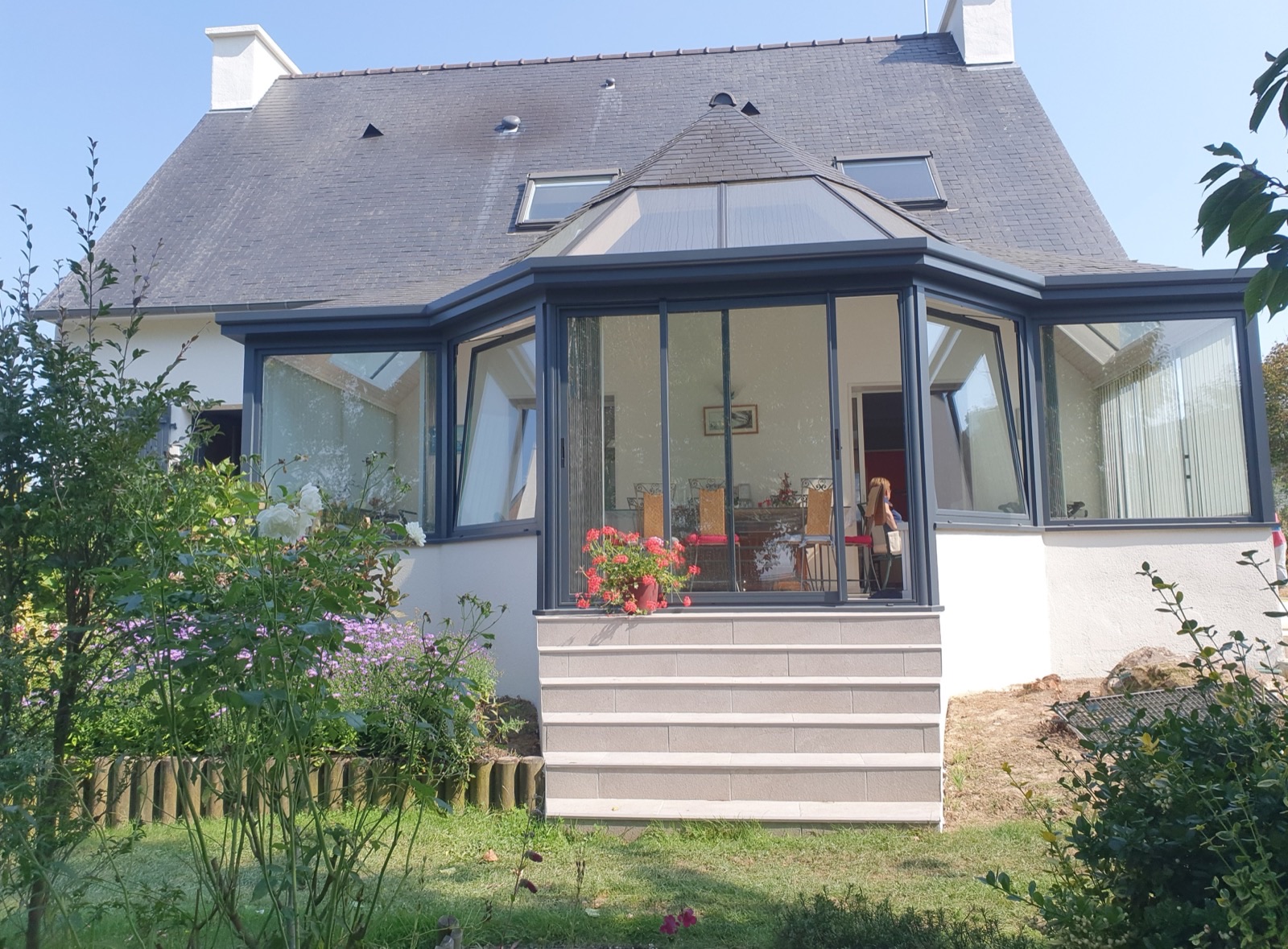 Maison avec véranda à structure grise et toiture en ardoise, jardin fleuri au premier plan