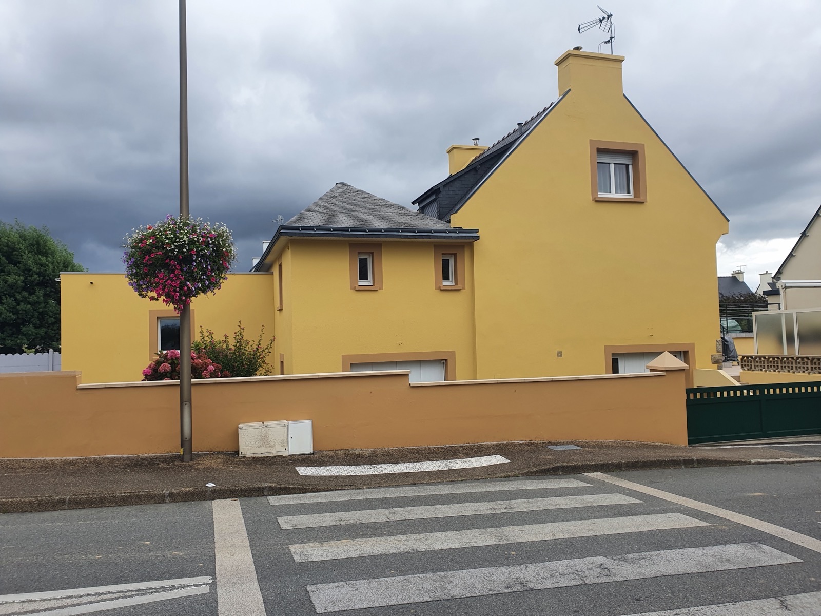 Maison et muret de clôture enduits en jaune, toiture en ardoise, sous un ciel nuageux
