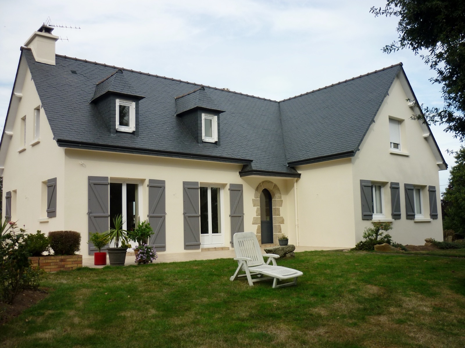 Maison néo-bretonne à façade claire, volets gris et lucarnes sur une toiture en ardoise