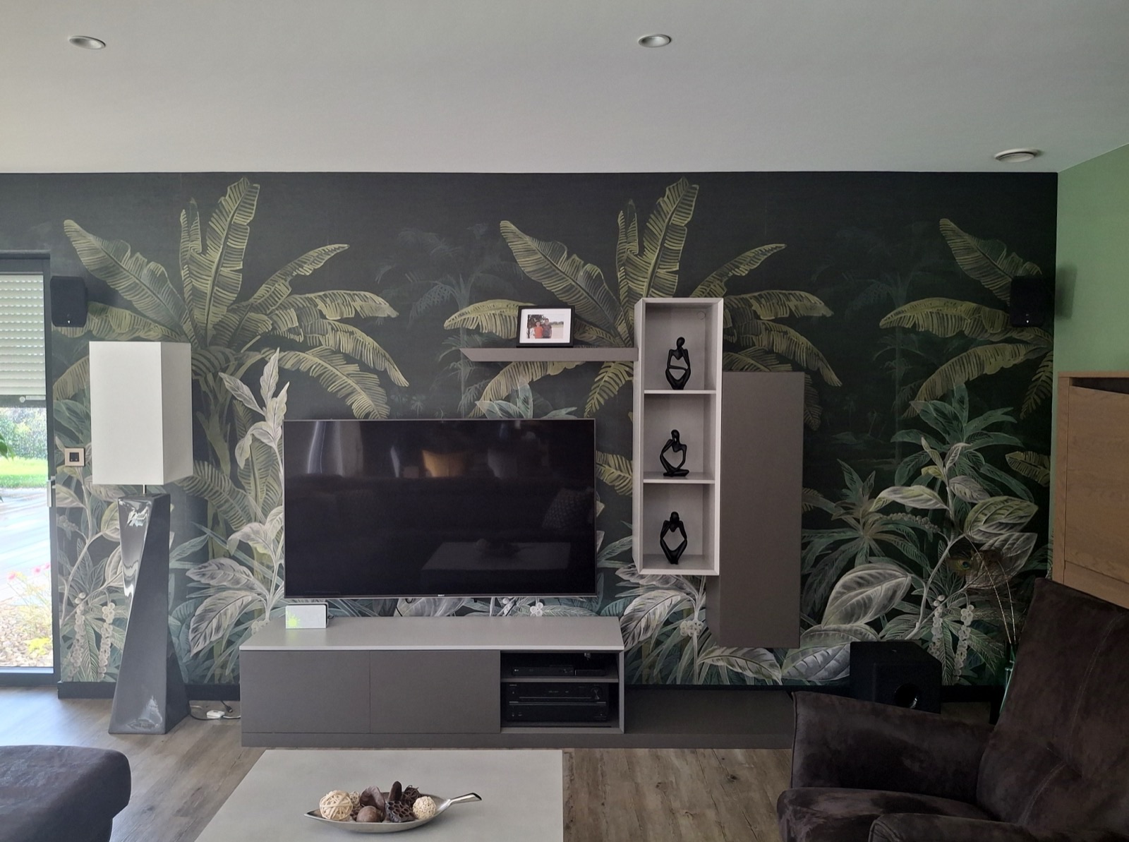 Salon avec papier peint panoramique jungle vert sombre derrière le meuble TV