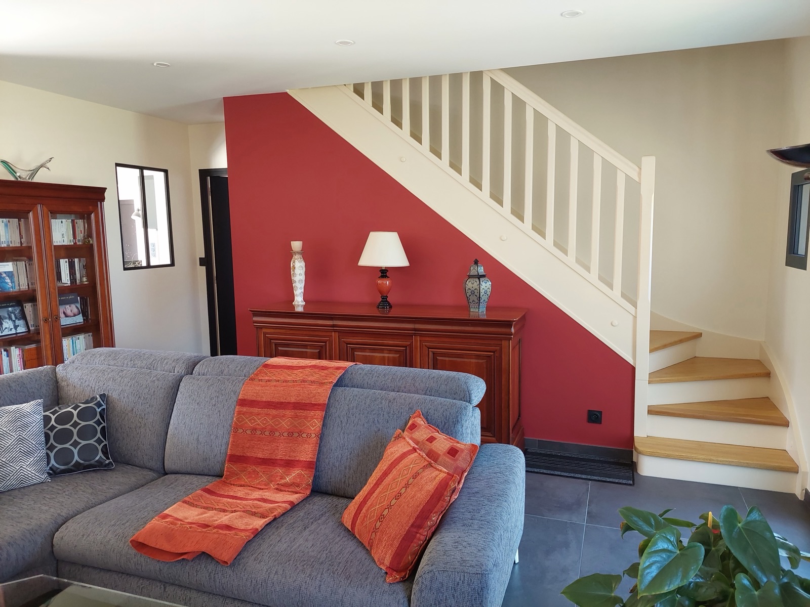 Mur peint en rouge sous un escalier blanc, canapé gris et buffet en bois
