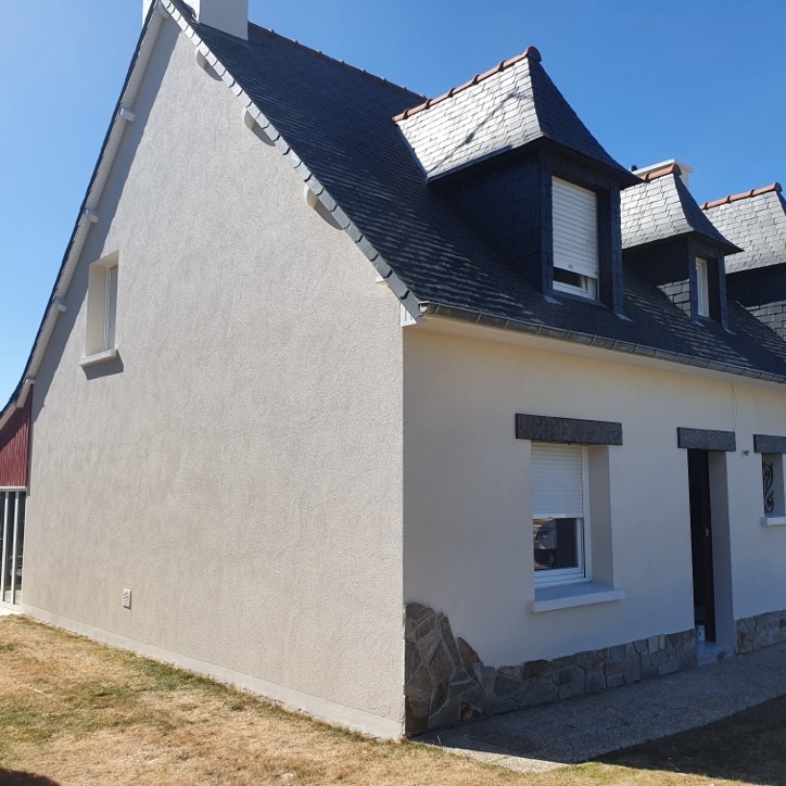 Maison néo-bretonne à façade enduite blanche, lucarnes et soubassement en pierre sous un ciel bleu