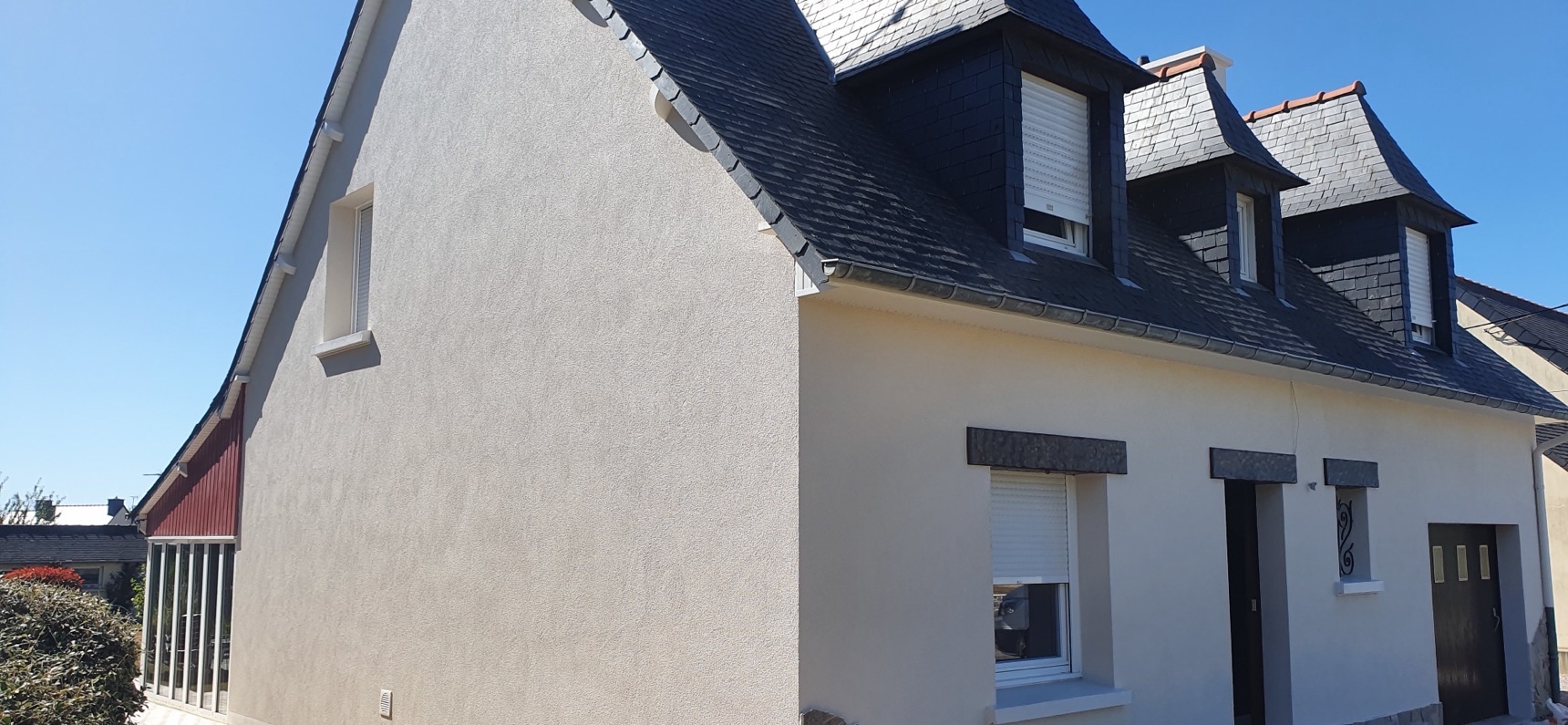 Maison néo-bretonne à façade enduite blanche, lucarnes et soubassement en pierre sous un ciel bleu