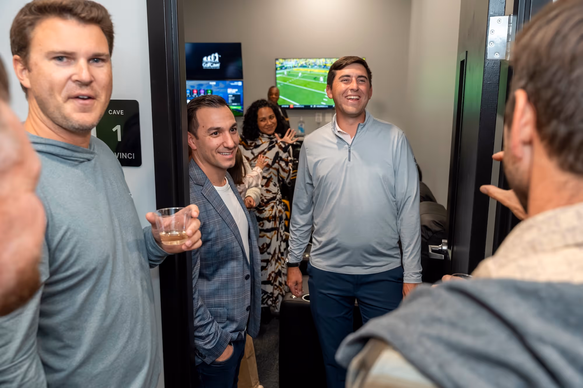 Man playing inside a golf simulator room at GolfCave with a group of people talking in the background at a golf party 