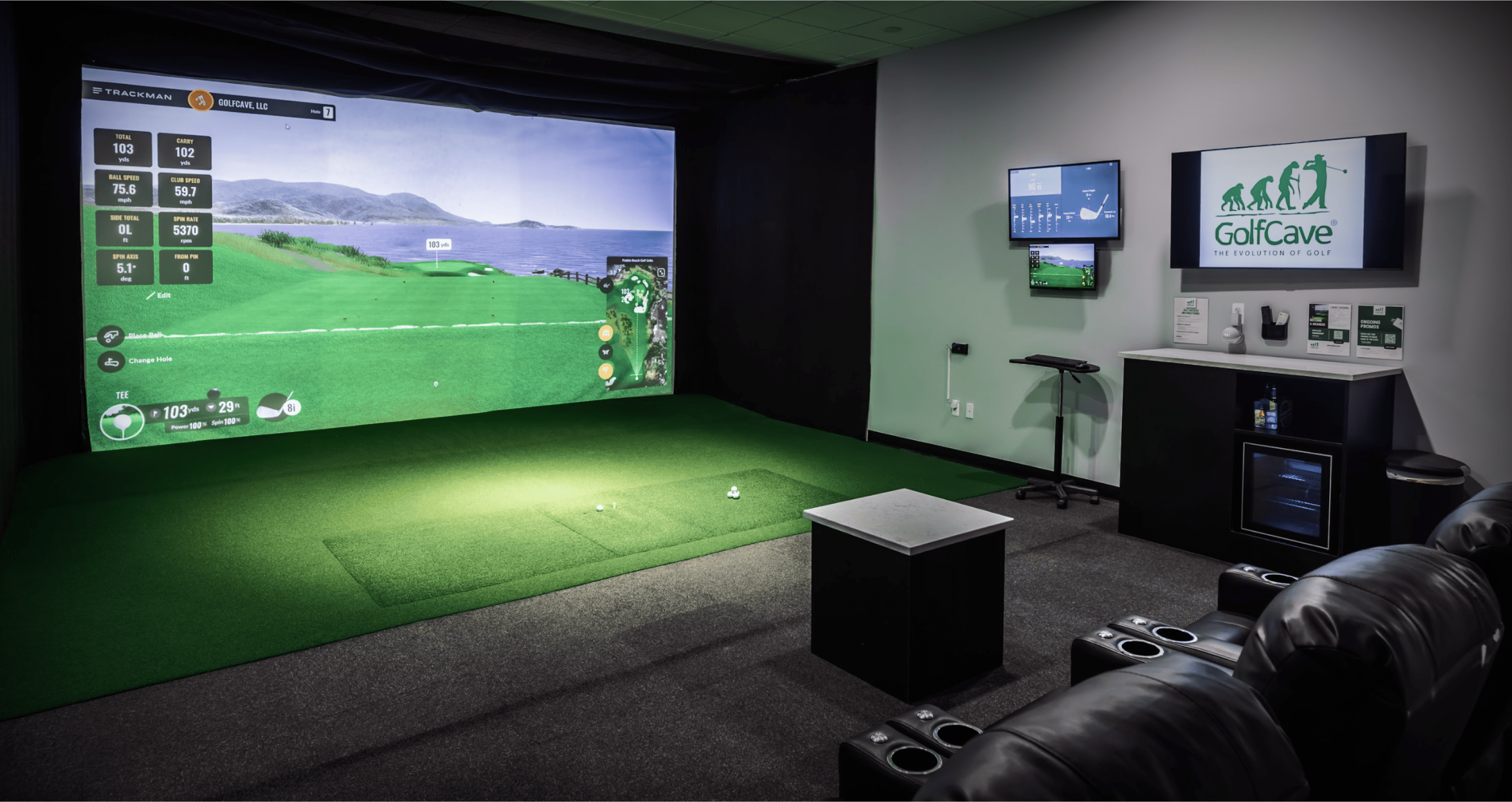 Panoramic view of an indoor golf simulator room showing the hitting area, dual monitors, counter top, and GolfCave on the TV
