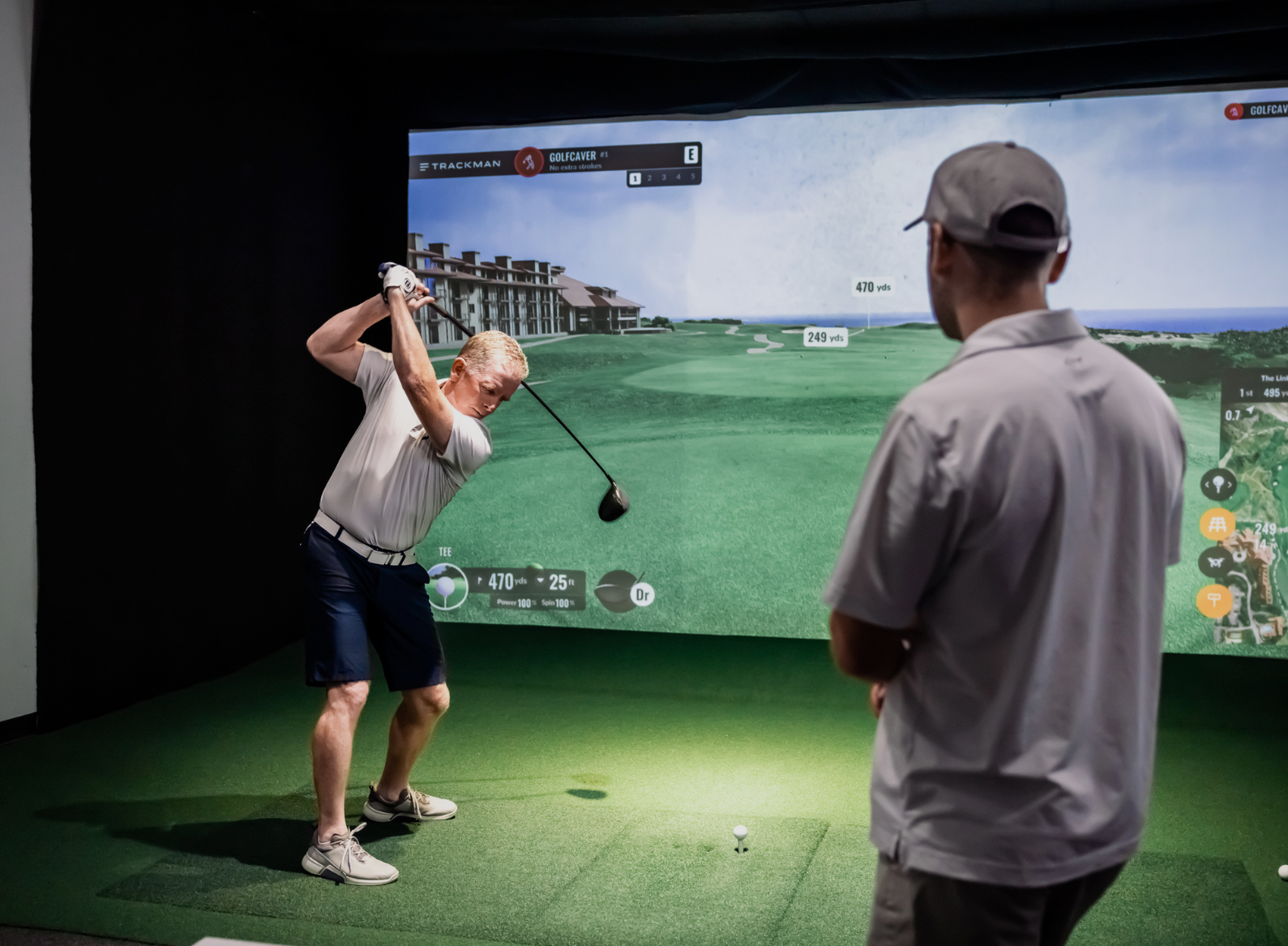 Two men inside an indoor golf simulator. One man is mid-swing with a driver while the other watches. A virtual golf course is projected on the screen showing distance markers and course details.