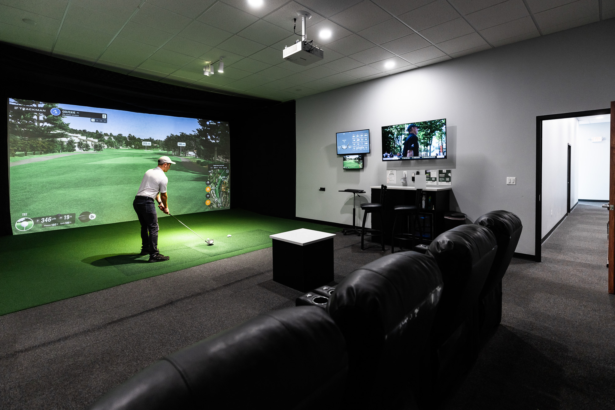 Panoramic view of an indoor golf simulator room showing the hitting area, dual monitors, counter top, and GolfCave on the TV