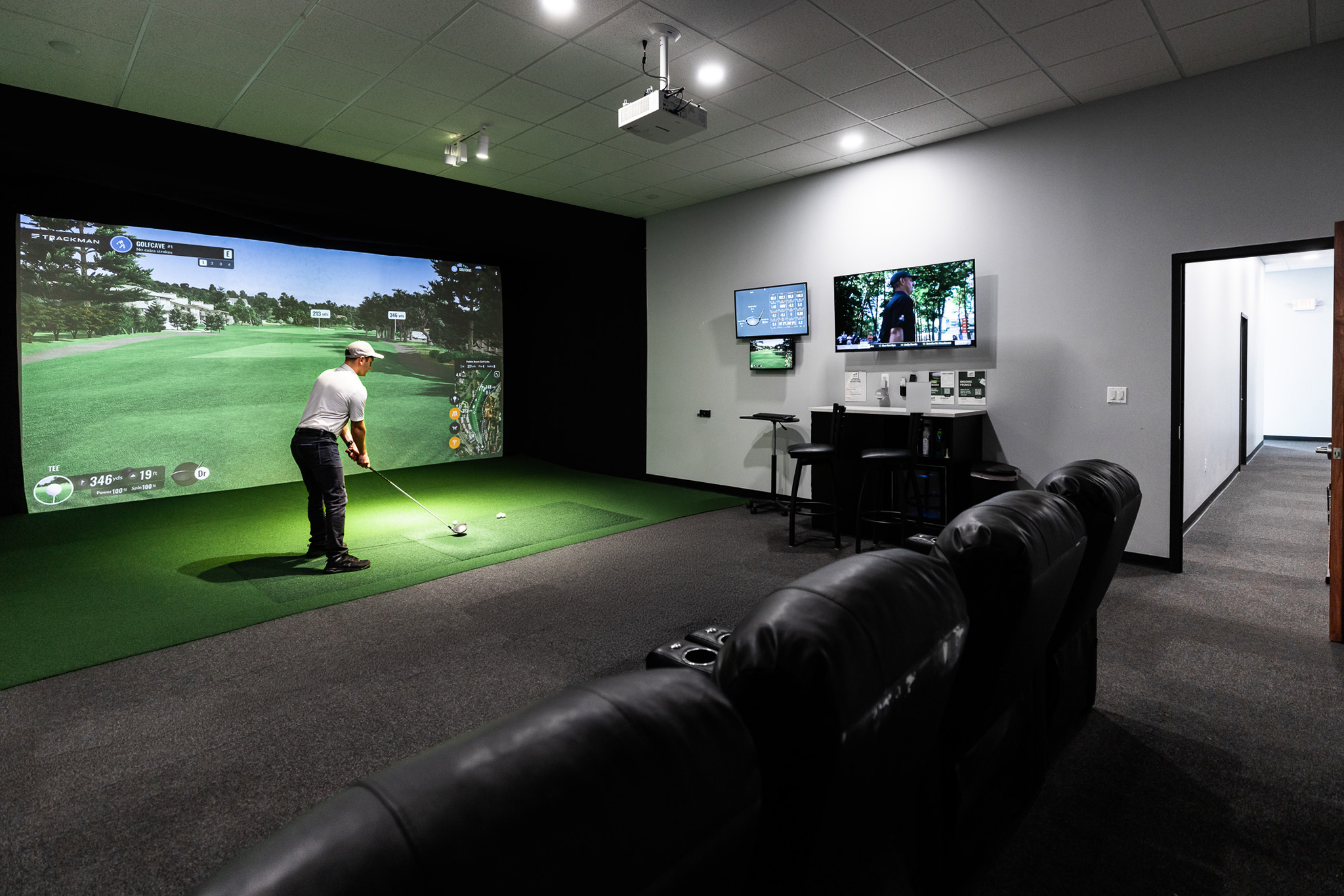 Panoramic view of an indoor golf simulator room showing the hitting area, dual monitors, counter top, and GolfCave on the TV