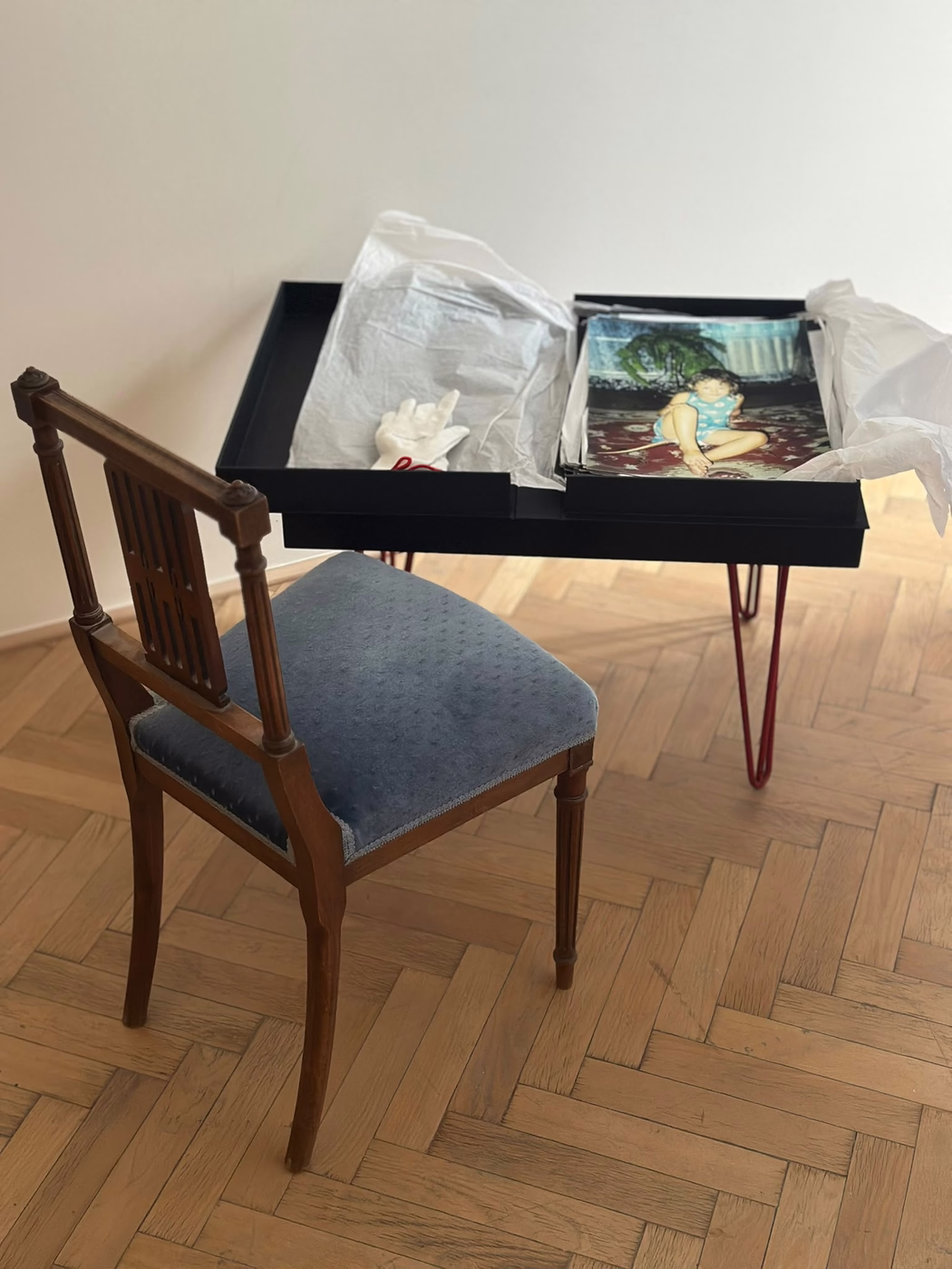 Dřevěná židle vedle stolu s černou krabicí obsahující fotografii dítěte a bílou rukavici.