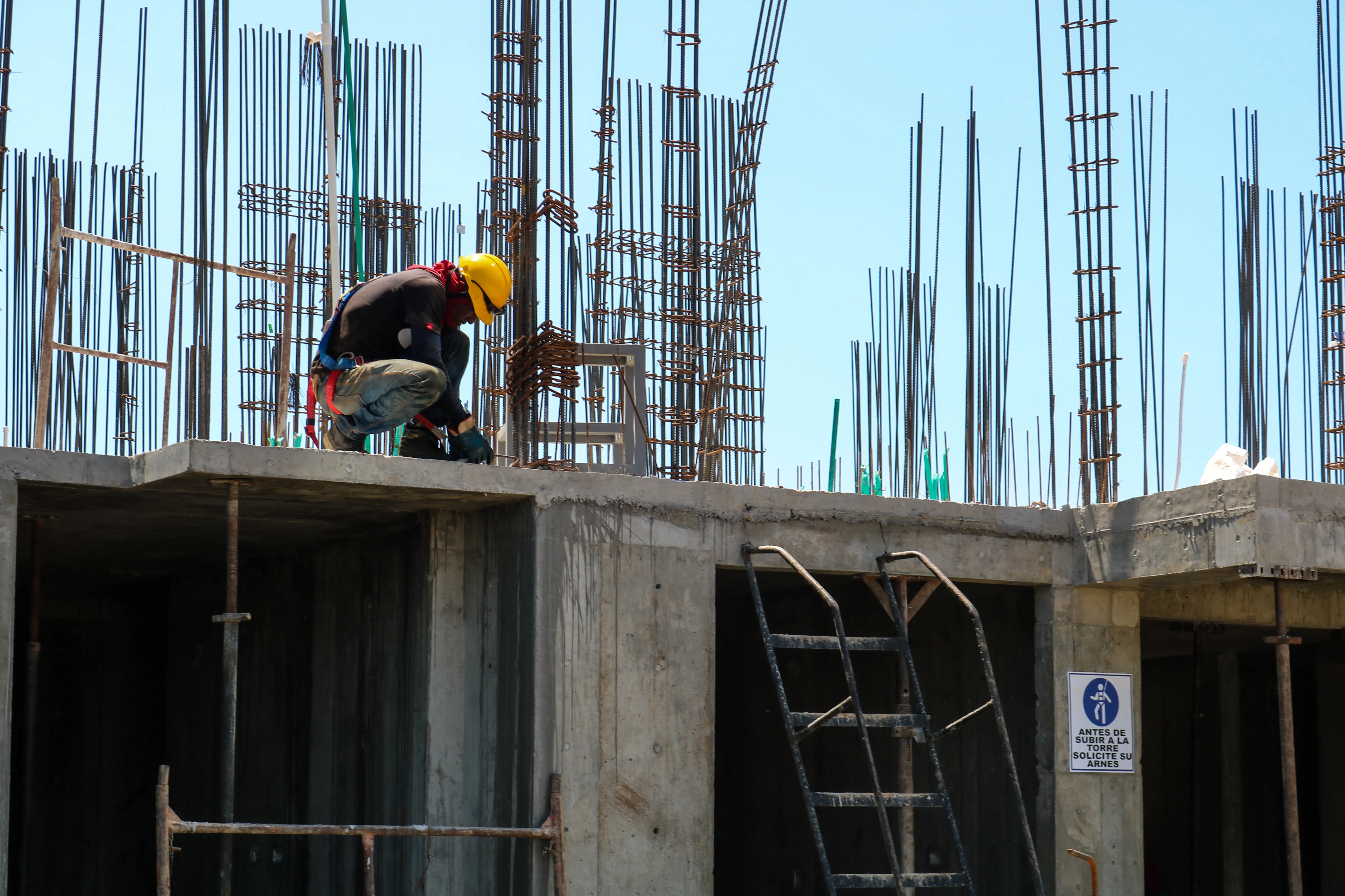 Construction worker wearing a yellow helmet and safety harness working on a building site with exposed rebar against a clear blue sky.