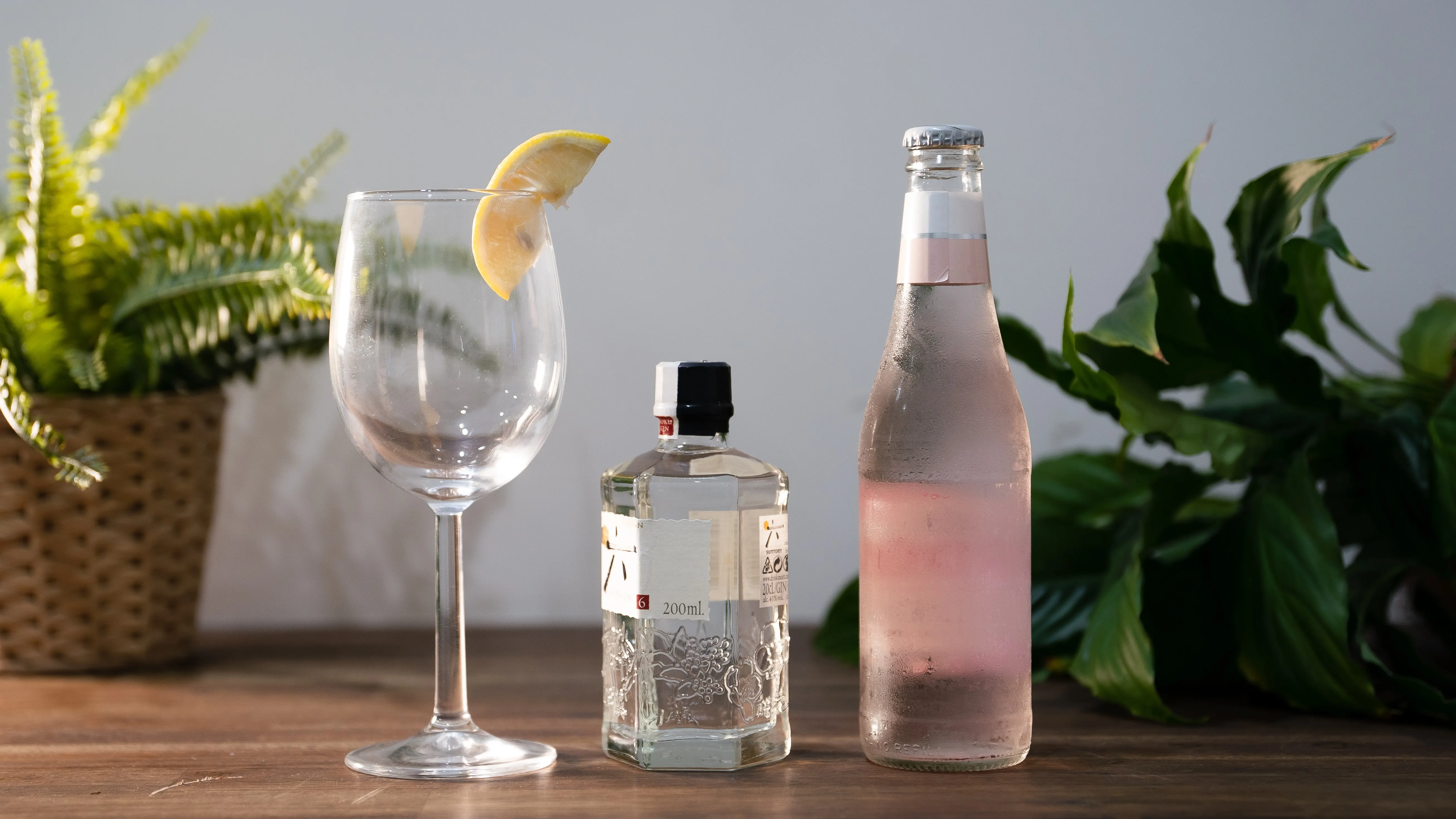 Empty wine glass with a lemon wedge on the rim, a small bottle of clear liquor, and a chilled pink bottle on a wooden surface with green plants in the background.